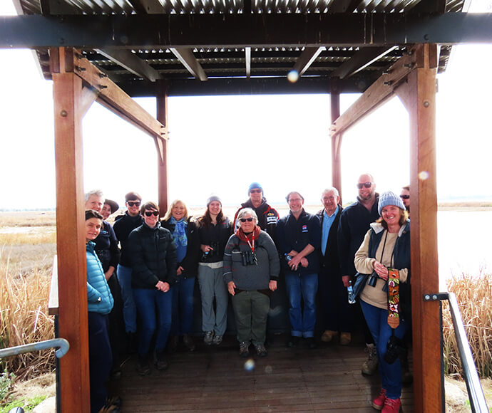 A group of individuals stands under a wooden shelter with a slatted roof, overlooking a marsh or wetland area. Most faces are obscured for privacy, except for one person in the center wearing sunglasses and a red scarf. The setting suggests an outdoor excursion or educational event related to the natural environment.