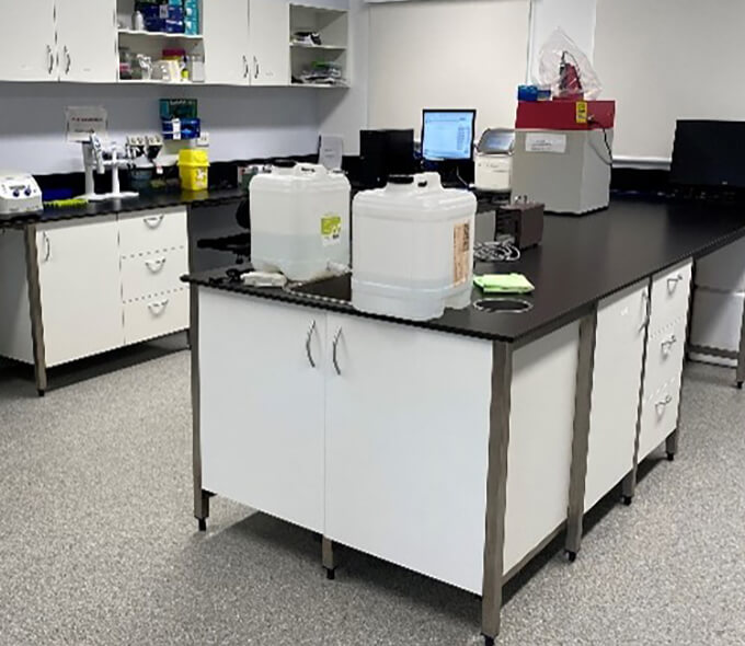 A modern laboratory with a black countertop and white cabinets, featuring various scientific equipment including a centrifuge, computer monitor, and containers labelled with blue tape.