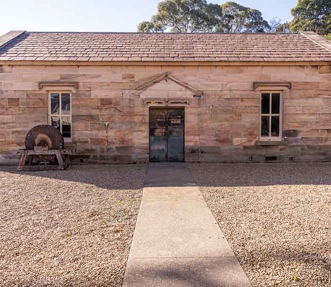 Single-story stone building with a shingled roof, central door, two windows, and old equipment on the left side