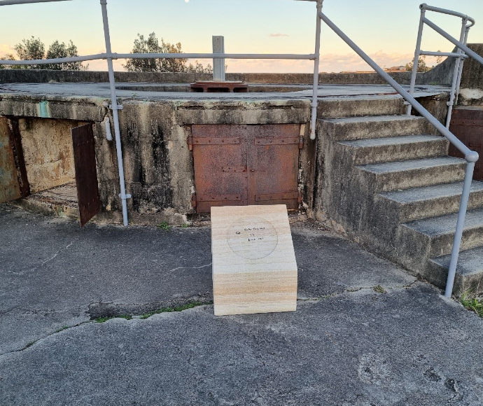 A light-coloured wooden plinth in front of a dark stone or cement historic structure, with a gently pink sunset behind