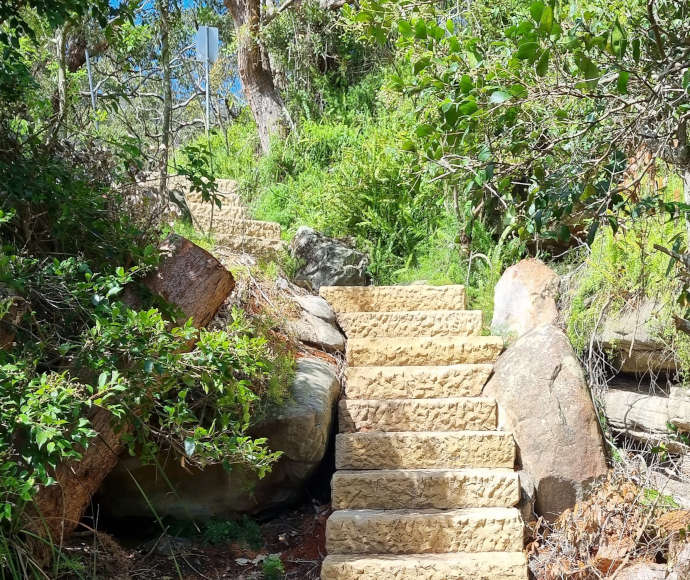 Sunlight hitting a roomy, rough-hewn sandstone staircase leading uphill through boulders and vivid green brush