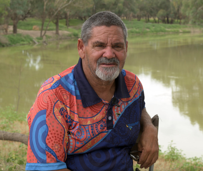 Murrawarri man Fred Hooper, leaning on a branch, beside still, brown water