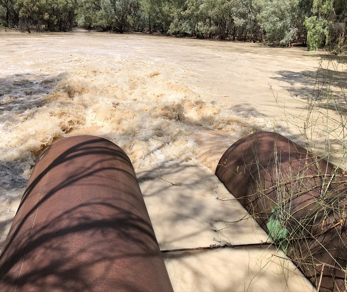 Two large cylindrical pipes discharging turbulent brown water into the Warrego River from Boera Dam, surrounded by natural vegetation.