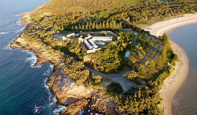 A bird's eye view of Trial Bay Gaol campground located in Arakoon National Park. The landscape features lush green trees with smooth sand lining the right side and gentle rocky cliffs lining the left side. There are a series of buildings and roads towards the centre of the landscape, with plenty of trees of various sizes taking up the majority of the space.