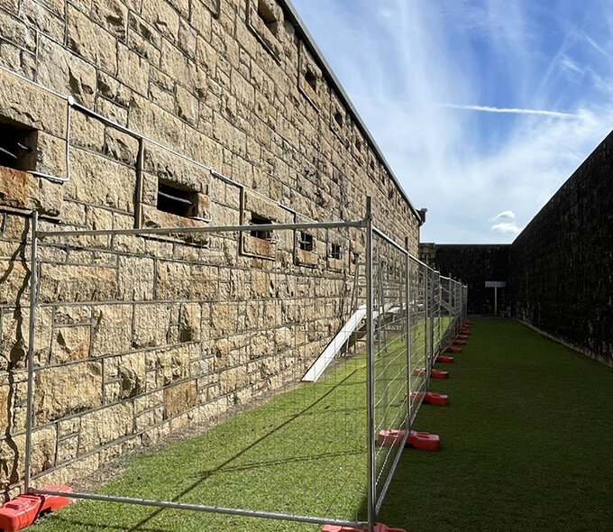 A stone wall with small windows and a metal fence on a grassy path under a blue sky