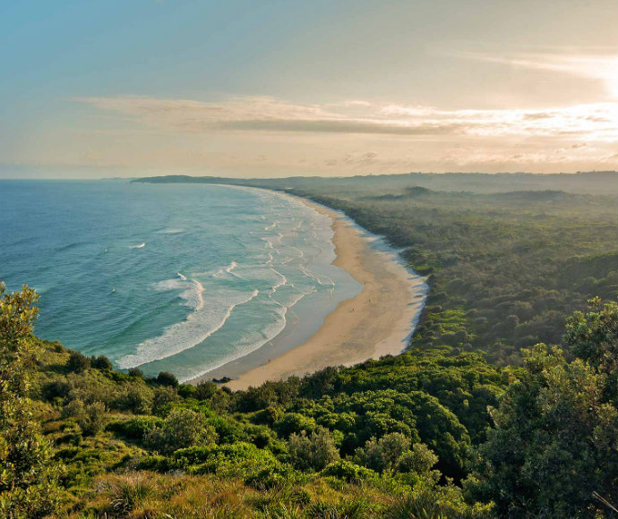 A beautiful view of Tallow Beach in Arakwal National Park, showcasing a long stretch of golden sand bordered by gentle waves from the deep blue ocean. Dense, green coastal vegetation lines the back of the beach, and Cape Byron can be seen in the distance. The sky is mostly clear with a few wispy clouds, adding to the serene and expansive feel of the coastline. The scene captures the pristine and natural beauty of Tallow Beach, a peaceful spot in the heart of New South Wales' coastal landscape.