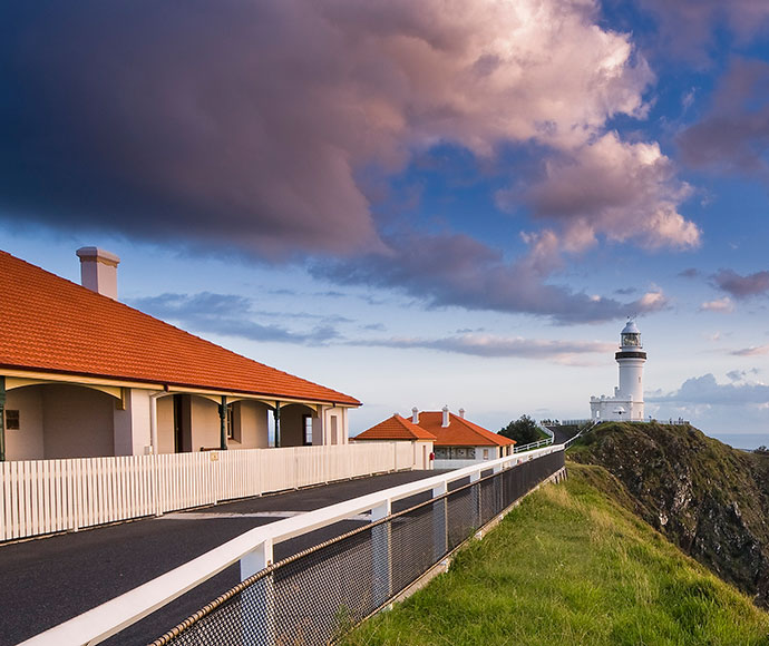 Cape Byron State Conservation Area Lighthouse: A scenic view of the Cape Byron Lighthouse in New South Wales, perched on a cliff surrounded by grassy slopes. The image features the lighthouse in the distance under a partly cloudy sky, with heritage-style buildings and a pathway leading toward it.