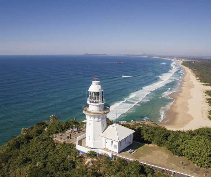 Aerial view of a lighthouse perched on a lush cliff, overlooking a vast ocean. Waves gently lap the sandy shore under a clear blue sky, conveying tranquillity.