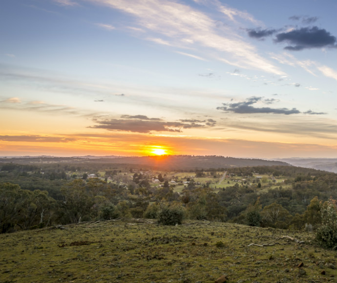 A scenic view of a countryside landscape at sunrise, seen from a hilltop lookout. The sun is rising on the horizon, casting a warm golden glow across the fields, trees, and small buildings scattered throughout the valley below. Soft clouds streak across the sky, painted with hues of orange, pink, and blue. The foreground includes grassy and rocky terrain, leading the eye toward the expansive, tranquil scene stretching into the distance.