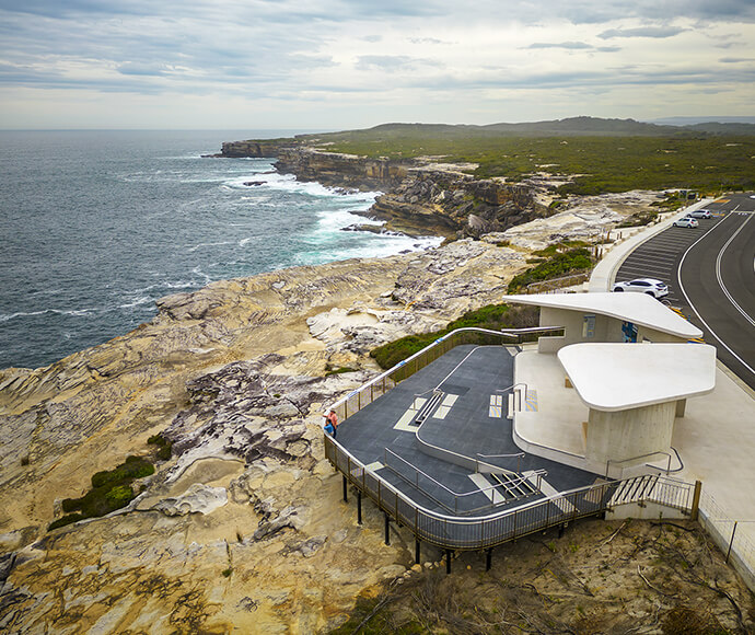 The image depicts the whale watching facilities at Cape Solander. There's a modern observation deck with seating and shade structures, built on rocky cliffs overlooking the ocean. A winding road with parked cars is visible to the right, and the coastline extends into the distance under a cloudy sky. The location is scenic and designed for whale watching.