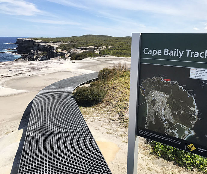 metal walkway curving through a coastal landscape with rocky cliffs and vegetation. A signboard titled "Cape Baily Track" displays a map of the area.