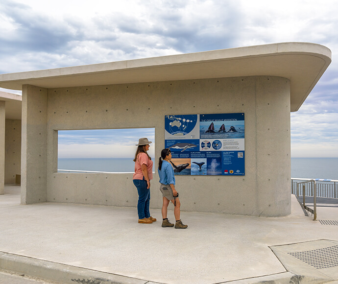 "Whale watching platform at Cape Solander, Kamay Botany Bay National Park. Two people stand by an informational display board on a concrete structure. The board contains text and images about whales and their migration. Behind the board is a large rectangular window overlooking the calm ocean under a cloudy sky.