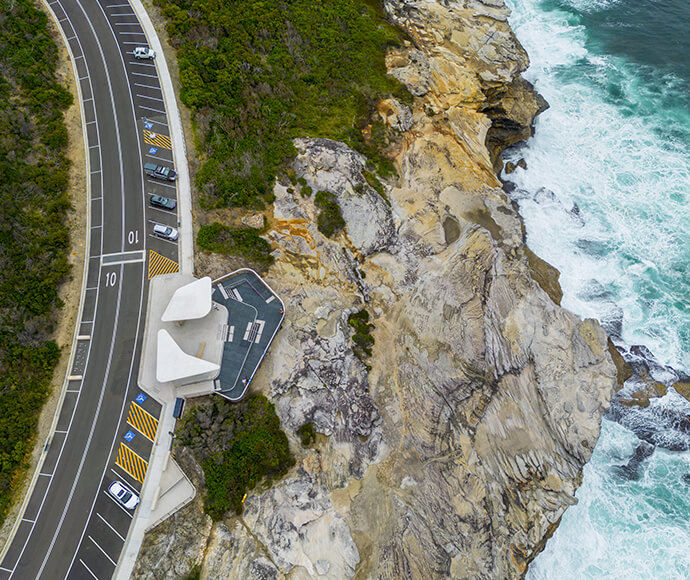 The image shows an aerial view of a whale watching platform at Cape Solander in Kamay Botany Bay National Park. The platform is situated on a rocky cliff overlooking the ocean, with waves crashing against the rocks below. To the left of the platform, there is a road with parked cars and a parking area. The platform itself is designed to provide a vantage point for visitors to observe whales in the ocean. The contrast between the rugged coastline and the structured platform makes this location interesting an