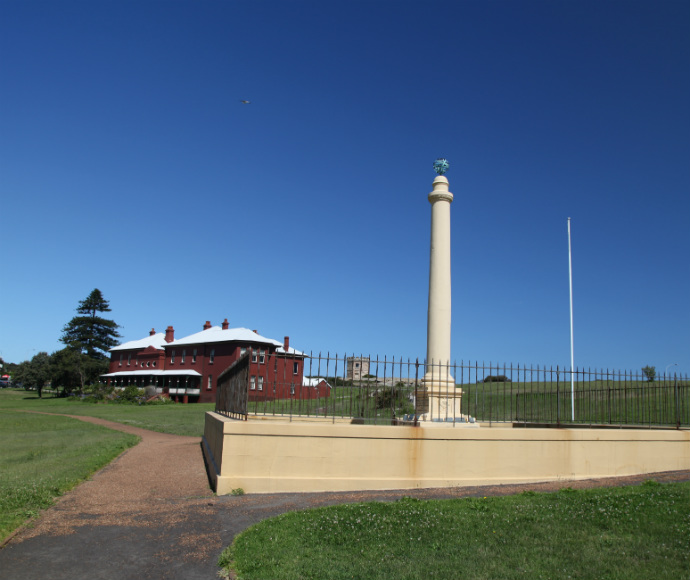 Kamay Botany Bay National Park Monument: A tall, cream-coloured historical column surrounded by a black iron fence, situated on a manicured lawn under a clear blue sky. In the background is a red colonial-style building with white roofs.