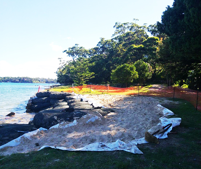 The image shows a coastal area in Bonnie Vale, part of Royal National Park, with a sandy beach on the left side. Large black sandbags are stacked along the shoreline, likely for erosion control. An orange plastic safety fence runs along the edge of the beach and into the grassy area, indicating a restricted or protected zone. The background features dense green trees and foliage, highlighting the natural surroundings alongside human intervention for environmental management.