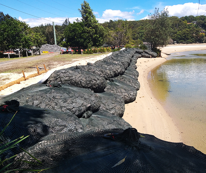 The image shows a shoreline in Bonnie Vale within the Royal National Park. In the foreground, large black mesh bags filled with rocks or sand are stacked along the edge of the water, forming a barrier to control erosion. The background features a grassy area and some construction equipment, indicating ongoing efforts to stabilize and protect the shoreline. Dense green trees and foliage can be seen further back, blending the natural landscape with human intervention for environmental management.