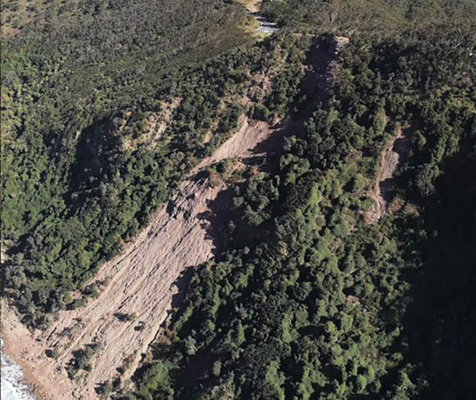 Aerial view of a landslip below Otford Lookout in 2022. The image shows a large section of the hillside where the vegetation has been displaced, revealing bare earth and debris. The surrounding area is densely forested, highlighting the contrast between the lush greenery and the exposed landslip area. The landslip appears to have affected a significant portion of the slope, indicating a substantial geological event.