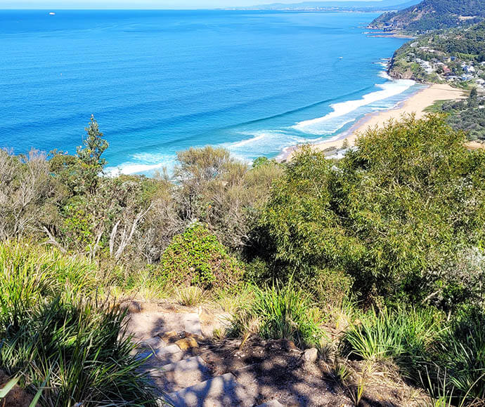 "Walkers enjoying the new safe pathway between Bald Hill and Stanwell Park. The image shows the pathway winding along the coastal landscape, with scenic views of the ocean, cliffs, and lush greenery. Walkers are seen taking in the breathtaking scenery while safely traversing the route.