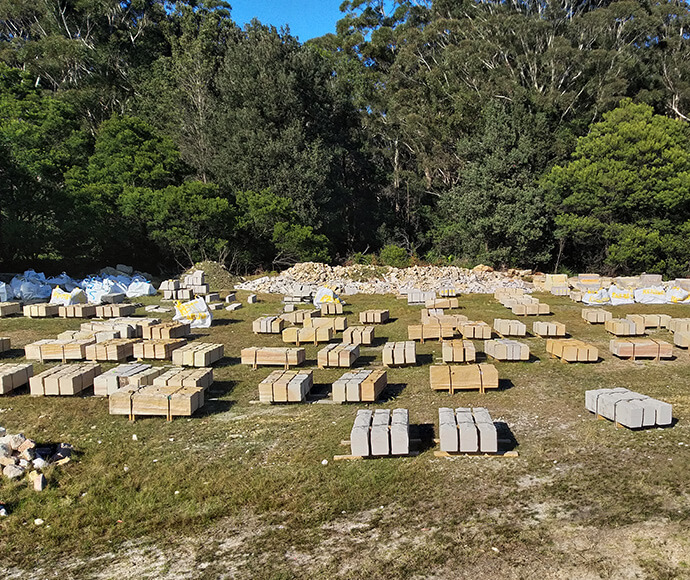 Outdoor construction storage site with neatly stacked concrete blocks on wooden pallets. Surrounding the site are piles of white bags and rubble. Dense trees and greenery frame the background, highlighting the organised storage of building materials in a natural setting