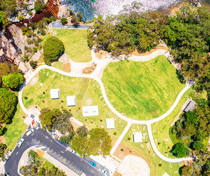 Aerial view of the Wattamolla picnic area in Royal National Park. The image showcases lush green lawns, pathways connecting various picnic shelters, and a parking area with several cars. The park is bordered by dense forest and a large body of water. Rocky terrain and dense trees surround the water, highlighting the well-maintained and picturesque recreational area.