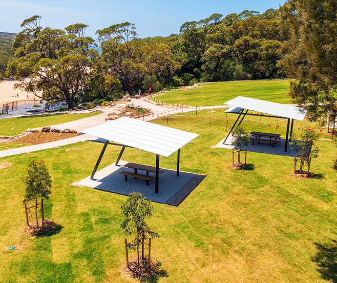 "New picnic shelters at Wattamolla in Royal National Park featuring metal roofs and wooden picnic tables on concrete slabs within a grassy area, surrounded by young trees and dense forest, with a sandy beach and body of water visible in the background.