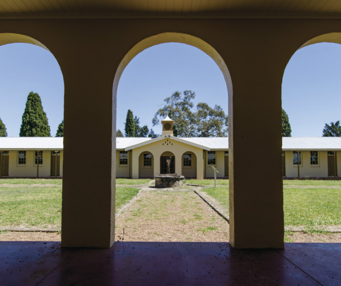 View through an arched walkway into a sunny courtyard with a central water feature. Surrounding buildings and trees evoke a serene, historic atmosphere.