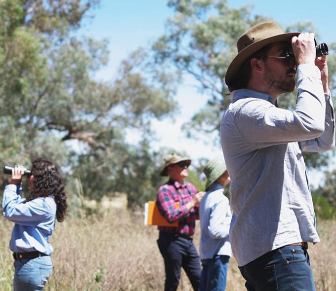 A group of people wearing hats and casual clothing surveying in a sunny, grassy area. They use binoculars, conveying a sense of focus and exploration.