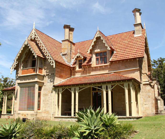 Victorian-style house with intricate gabled roof, patterned red tiles, and ornate woodwork. Surrounded by lush greenery under a clear blue sky.