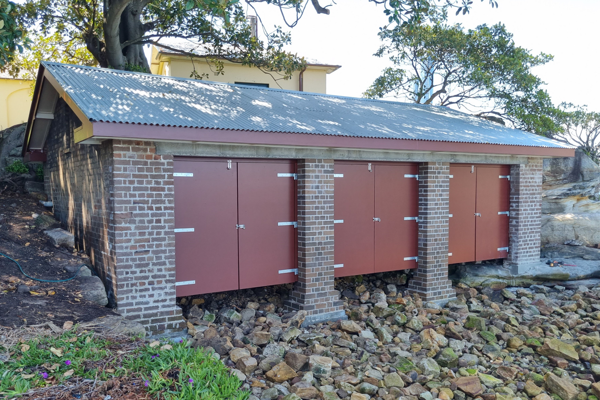 A brick boat shed with three closed red doors, and a corrugated iron roof.