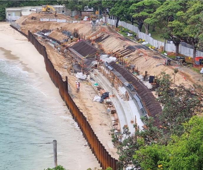 High view of a small inlet with pale green-blue waters lapping up to a small sandy beach full of construction works, leading up to a thick green treeline