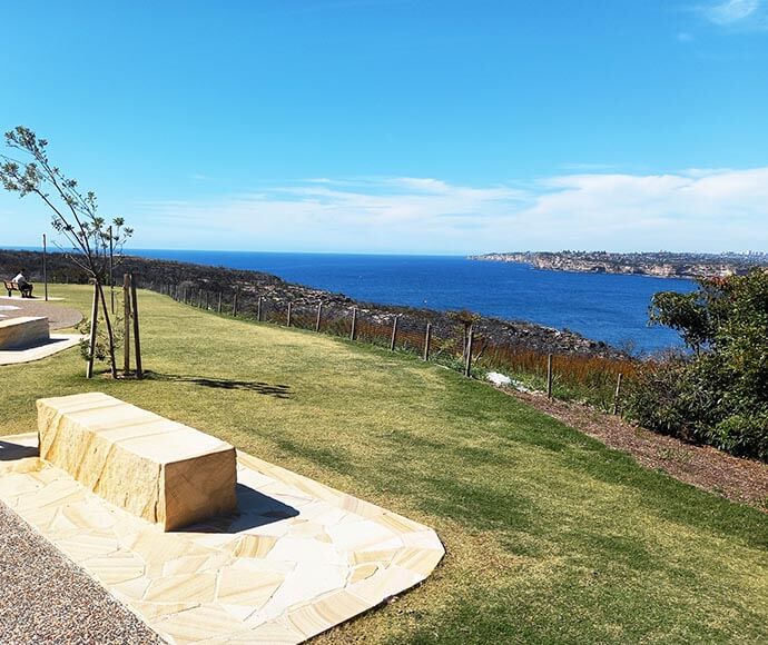 Completed landscaping at North Head, Sydney Harbour National Park, featuring a stone bench on a paved area, surrounded by native vegetation and scenic harbour views.