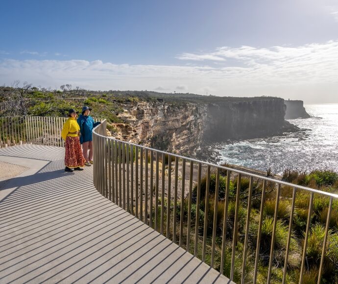 Yiningma Lookout at North Head, Sydney Harbour National Park, featuring a paved pathway leading to a scenic viewpoint with expansive ocean views, a couple enjoying the harbour view, and surrounding native vegetation.