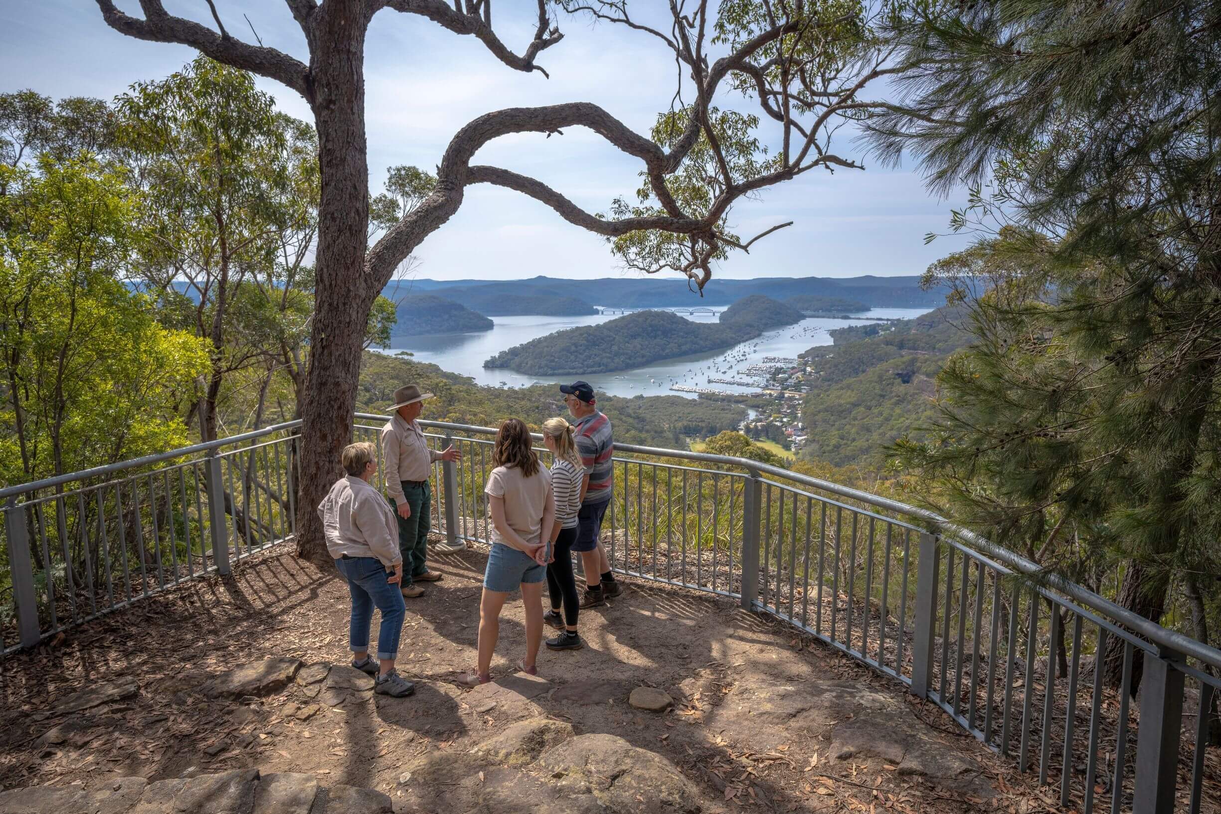 A group of five people stand on a lookout platform surrounded by trees, overlooking a scenic view of a river winding through a hilly landscape with boats and houses visible below.