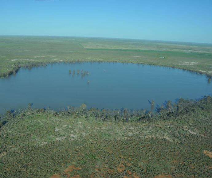 The image shows an aerial view of a large, circular body of water surrounded by a vast expanse of flat, green land. The water body appears to be a lake or a pond, with some trees or vegetation emerging from the water near the center. The surrounding land is covered with sparse vegetation, and the horizon is visible in the distance under a clear blue sky.