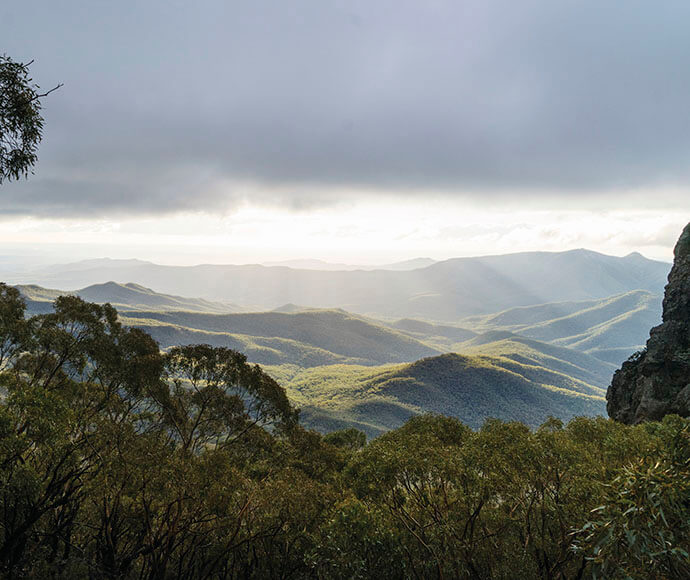 The image shows a scenic view of a mountainous landscape. The foreground features trees with dense foliage, while the background reveals a series of rolling hills and mountains extending into the distance. The sky is partly cloudy, with sunlight breaking through the clouds, casting rays of light onto the hills below. This interplay of light and shadow highlights the contours of the landscape, creating a visually striking and serene scene.