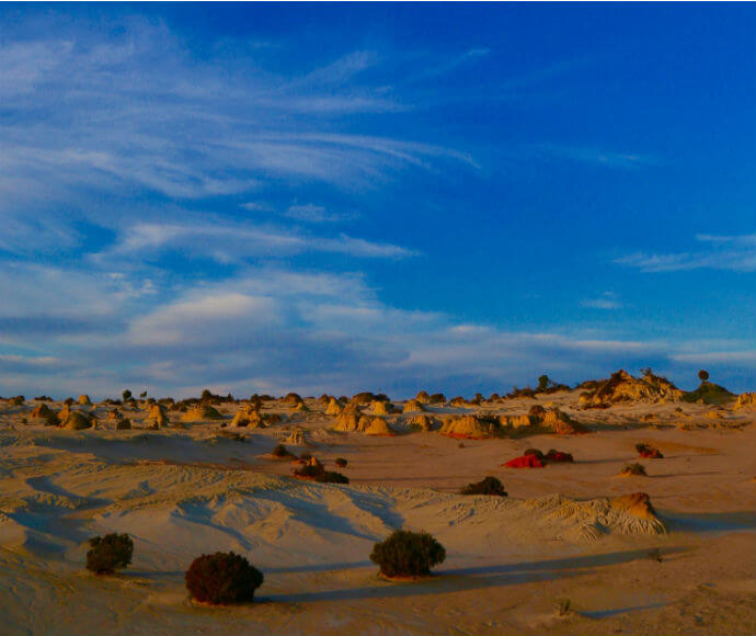 "A landscape view of the Mungo Lunette, also known as the Walls of China, featuring sand dunes and sparse vegetation under a blue sky with scattered clouds. The unique geological formations and the contrast between the sand and the sky create a striking and picturesque scene.