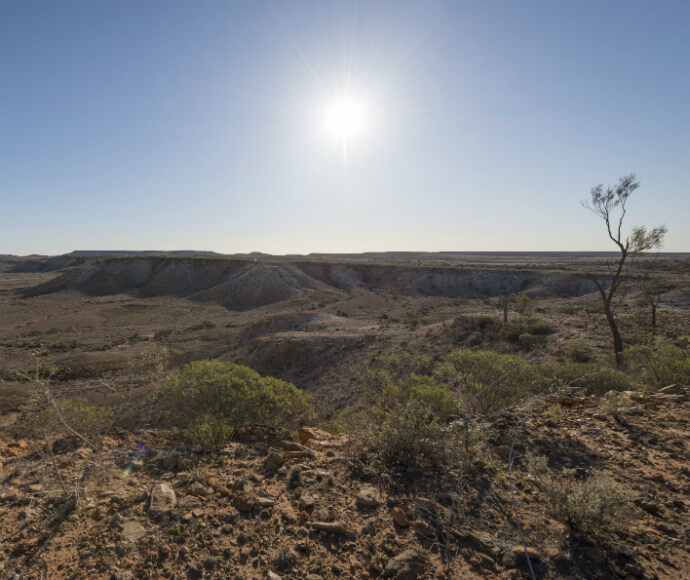 A vast, arid landscape under a clear blue sky with the sun shining brightly. The terrain is rugged with sparse vegetation, including small bushes and a few trees. The horizon stretches far into the distance, highlighting the remote and expansive nature of the Jump Up walking track in the northern part of Sturt National Park.