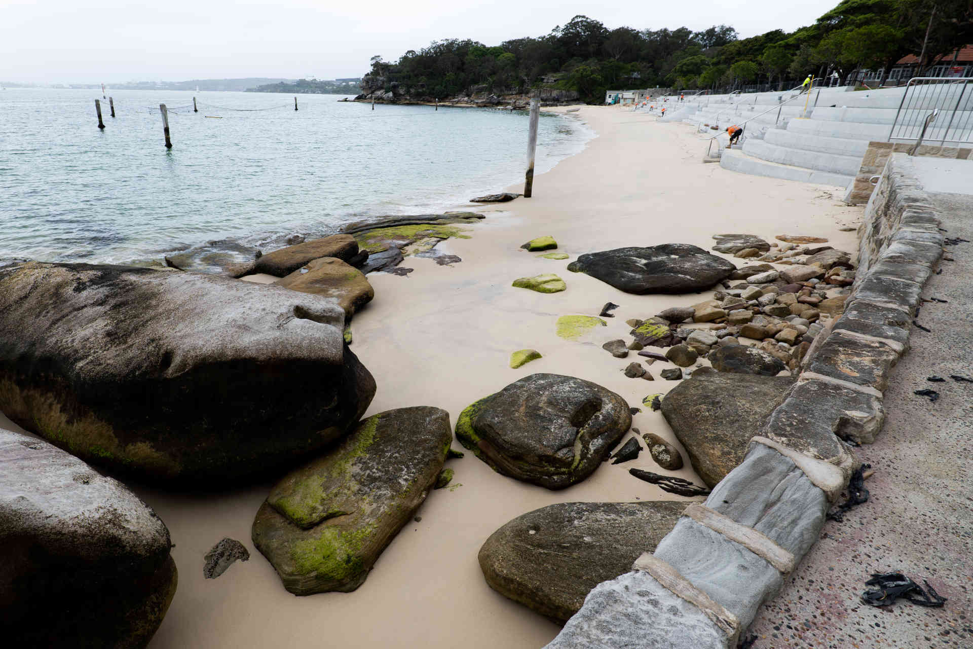 A photograph of a small bay on a bleary day viewed from a far corner of its narrow sandy beach, with fresh concrete bleachers visible on one side and a thick dark line of foliage at the far side