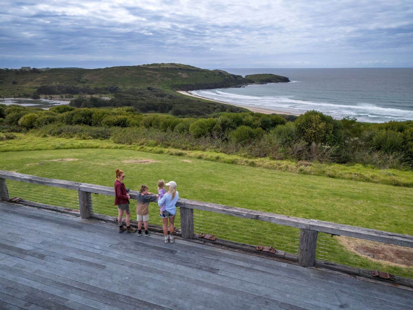 Four people stand on a wooden deck overlooking a scenic coastal landscape with green hills, a sandy beach, and the ocean. The sky is partly cloudy.