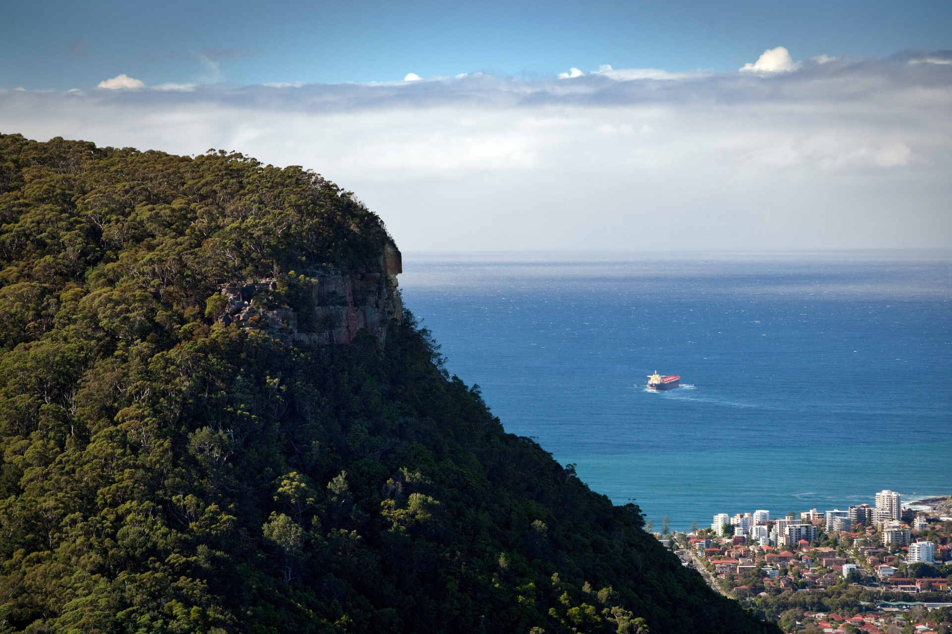 A thickly treed escarpment juts up on the left with a a cornflower blue ocean in the background and at its base, a city by the sea which looks like a toy city in the wake of the escarpment