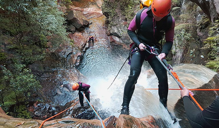 Two people abseiling down a waterfall, one has just started to descend, and the other is almost at the base.