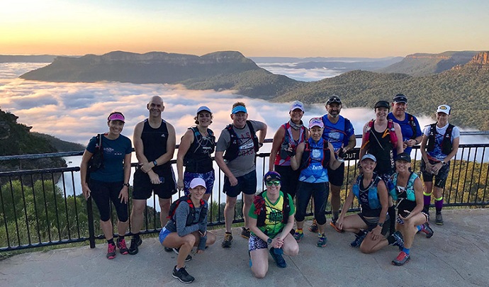 A group of runners standing at a scenic overlook. In the background, the majestic Blue Mountains rise, and below them, a layer of clouds stretches out.