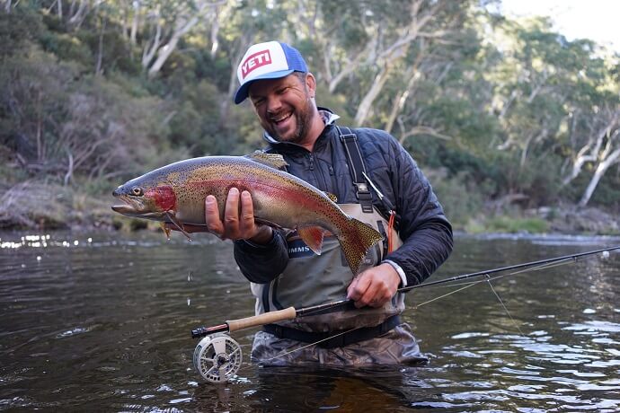 A man in outdoor clothing holding a large fish in front of him, standing in a river with a fishing rod and reel at his side.