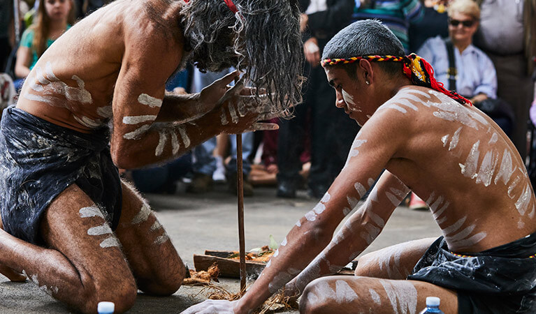 Two Aboriginal men crouched down while starting a fire. They are painted in traditional custom while a crowd of people watch.