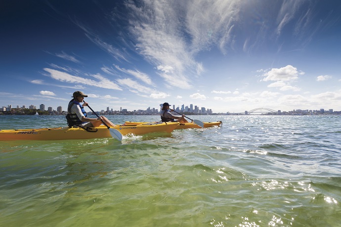 Two individuals kayaking on a clear day with a city skyline and bridge in the background, under a blue sky with wispy clouds.