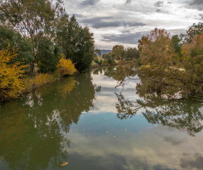 A peaceful scene showing a small, still river with green, yellow and brown foliage at its banks, grey clouds above and a silhouetted mountain in the distance