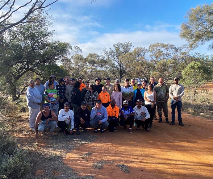 Group photo of multiple individuals outdoors with trees and shrubs in the background on dry, dusty ground.