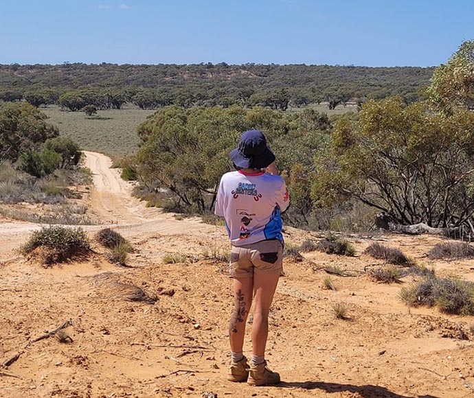 Ranger in bucket hat and shorts, back to camera,  staring down dusty road