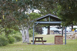 Picnic area with a wooden picnic table under a small open shelter with a gabled roof, surrounded by grassy grounds, scattered leaves, and trees. A white vehicle and another structure are partially visible in the background.