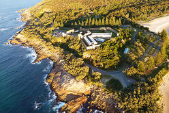 This image shows an aerial view of Trial Bay Gaol campground. The historic gaol building is central, surrounded by greenery and bordered by the ocean and beach. The landscape features winding roads, dense forest, and rocky shorelines. The viewpoint offers a comprehensive look at the natural beauty and historical significance of the coastal location.